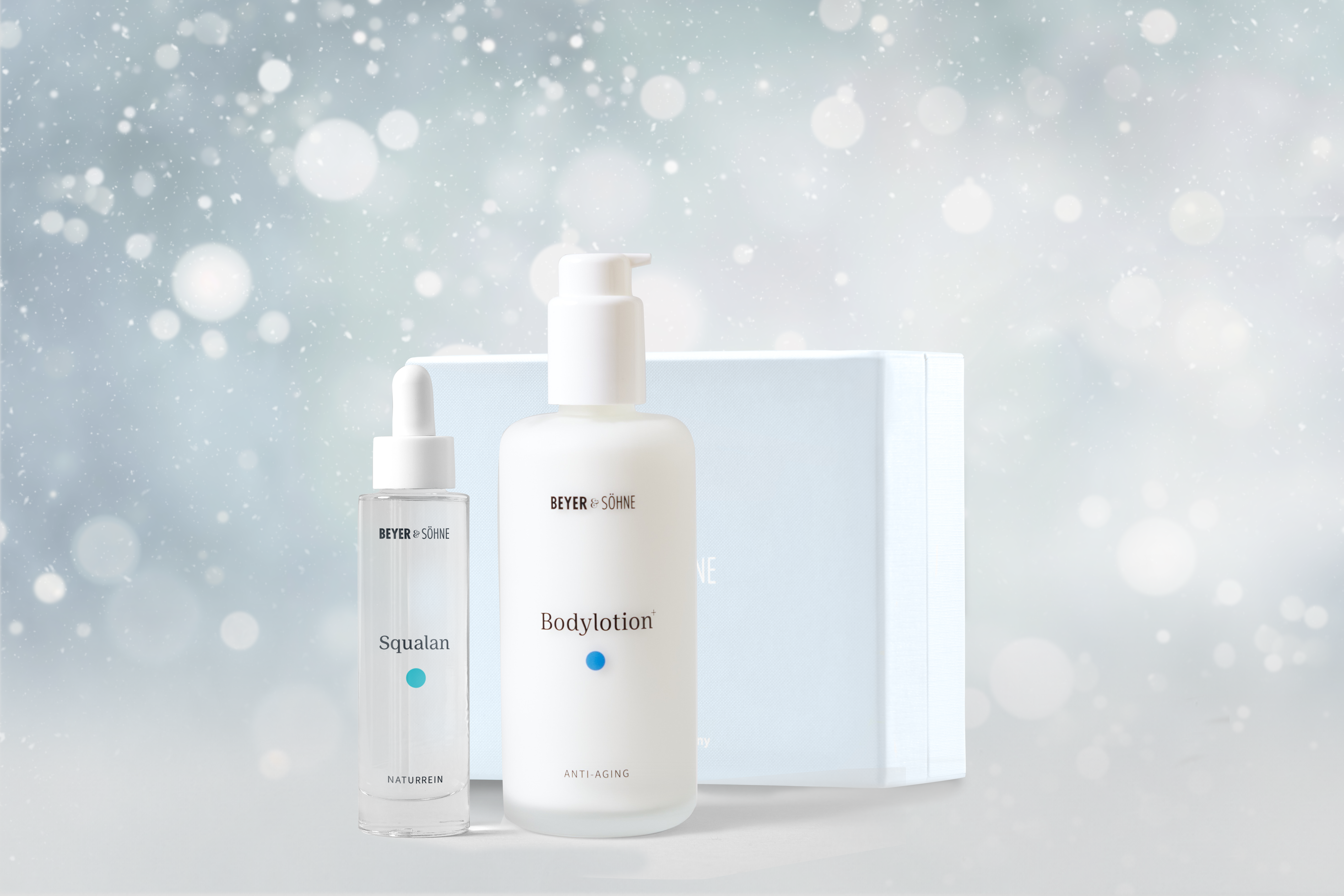 Bodylotion und Squalan in hochwertigen Glasspendern von Beyer & Söhne vor himmelblauer Geschenkbox und weihnachtlich gestimmten Hintergrund mit Schneeflocken.